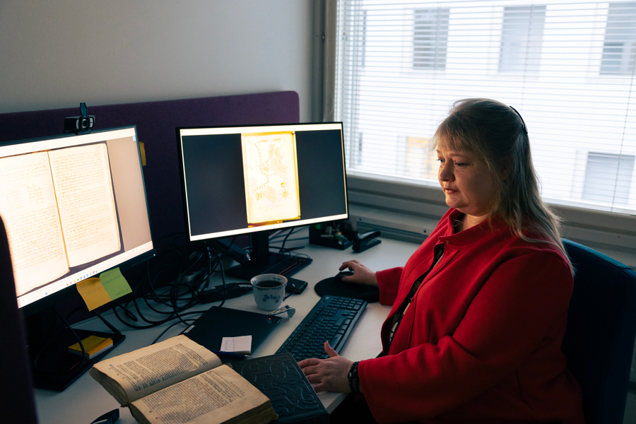 Raisa Toivo tietokoneen ääressä, pöydällä avoinna vanha kirja.