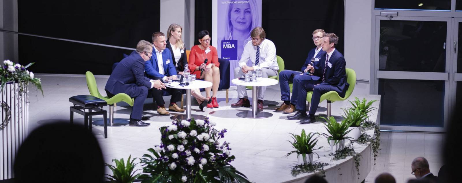 Johtajuussymposiumin panelisteja lavalla, yleisön siluetteja etualalla.