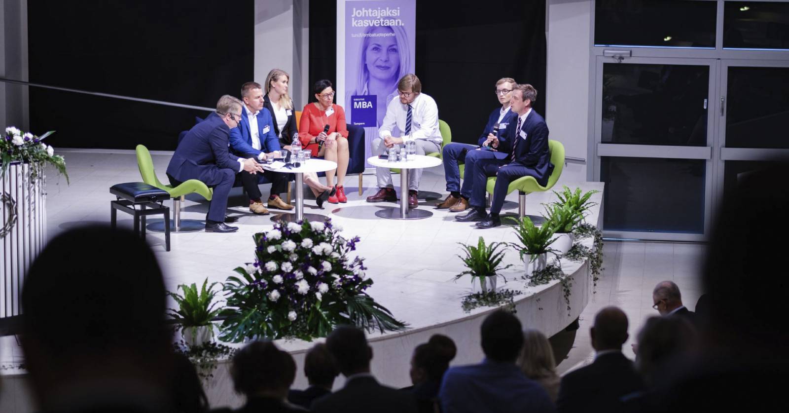 Johtajuussymposiumin panelisteja lavalla, yleisön siluetteja etualalla.