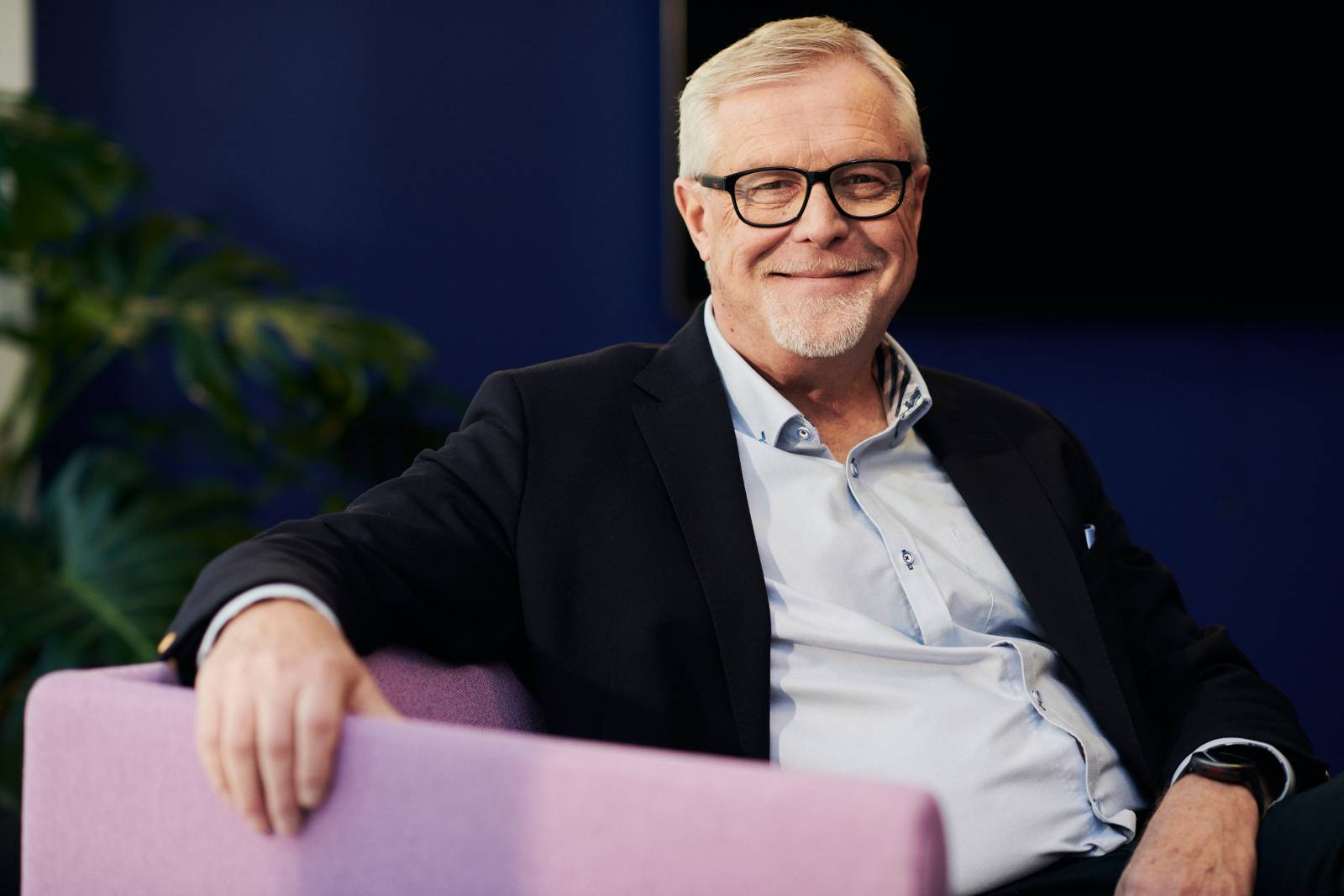 Harmaahiuksinen ja silmälasipäinen mies istuu nojatuolissa, kasvot kohti kameraa ja hymyilee