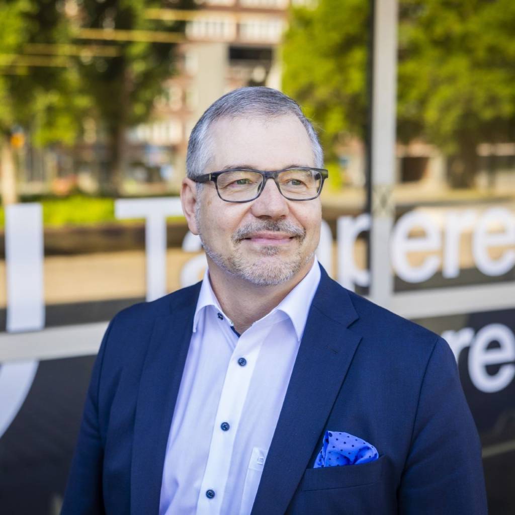 Rehtorilla on harmaat hiukset ja mustapokaiset silmälasit. Hän seisoo lasiseinän edessä, jossa on Tampereen yliopiston logo. Hämäläisellä on valkoinen kaulupaita ja tummansininen pikkutakki. Hän hymyilee ja katsoo viistosti oikealle, ei suoraan kameraan.