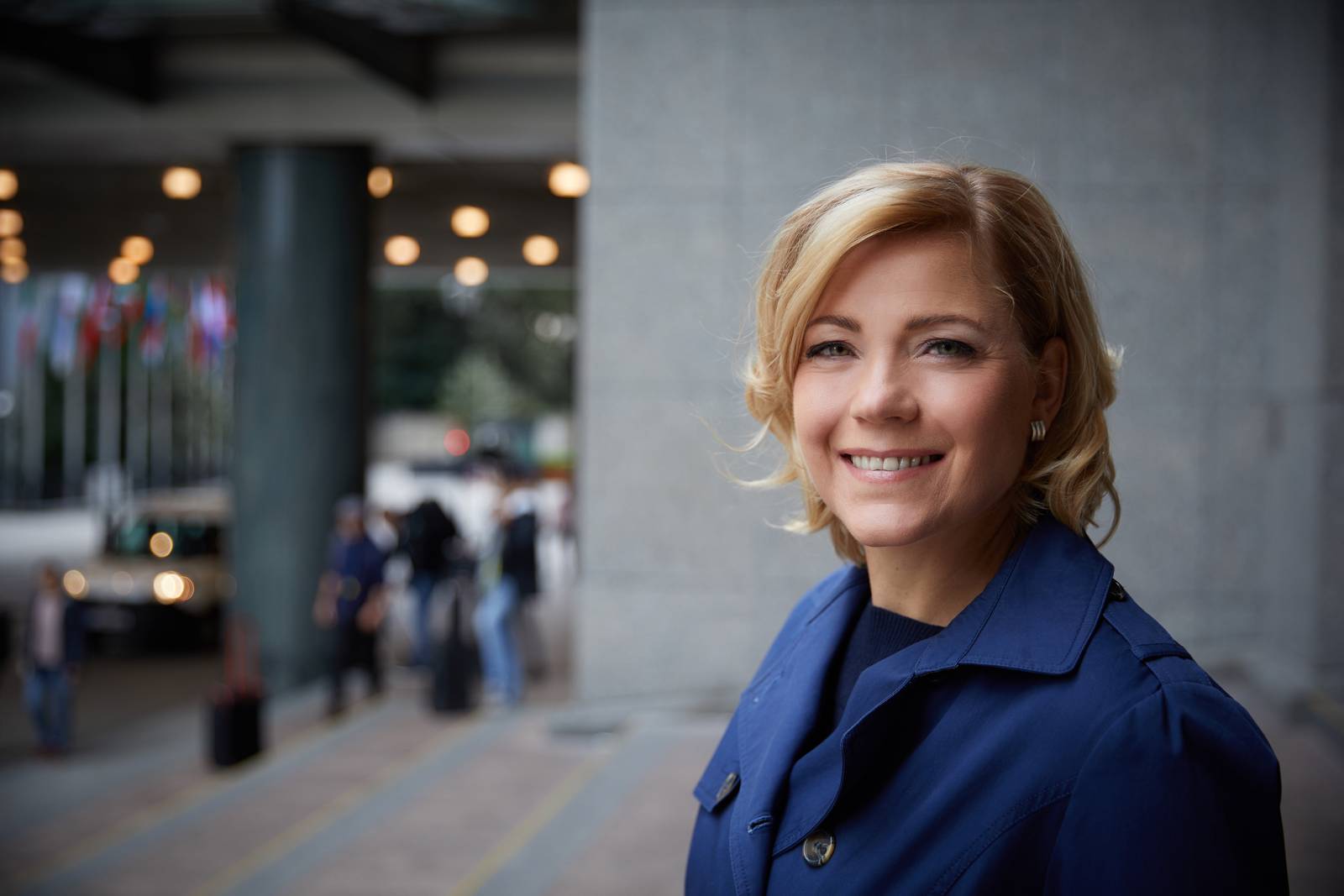 Henna Virkkunen, jolla on vaaleat, polkkamittaiset hiukset, katsoo kameraan ja hymyilee. Hänellä on yllään musta paita ja tummansininen jakkutakki.