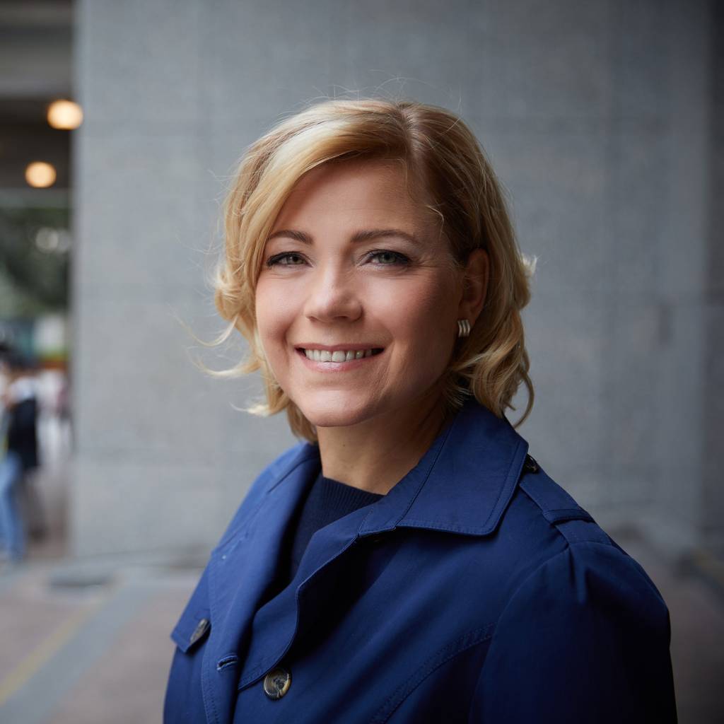 Henna Virkkunen, jolla on vaaleat, polkkamittaiset hiukset, katsoo kameraan ja hymyilee. Hänellä on yllään musta paita ja tummansininen jakkutakki.