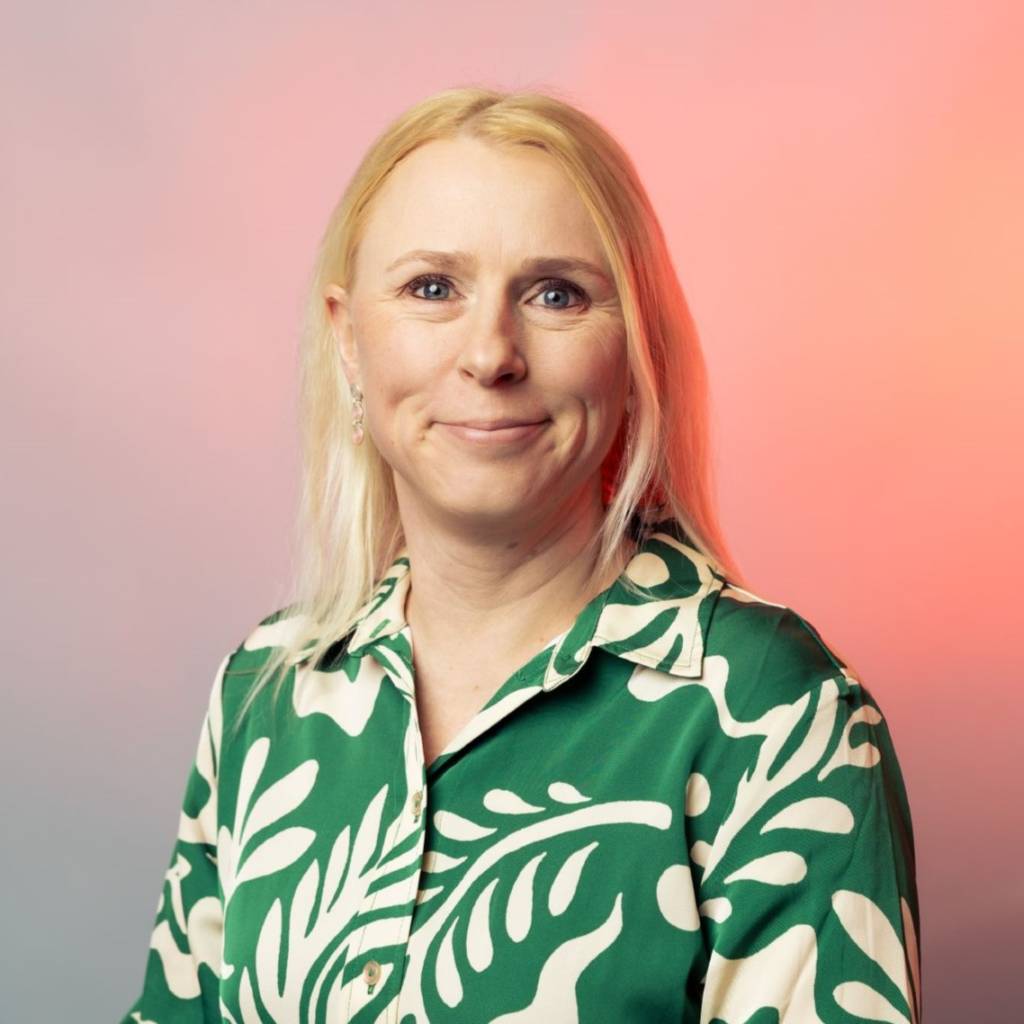 Elina Kestilä-Kekkonen, jolla on vaaleat, pitkät hiukset, katsoo kameraan ja hymyilee. Hänellä on vihreä-valkoinen kirjava napitettu pusero yllään.