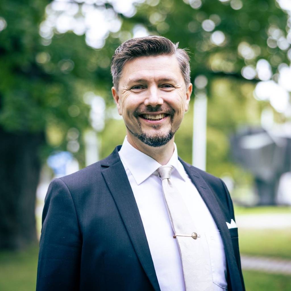 Thomas Olsson katsoo kameraan ja hymyilee. Taustalla näkyy vihreitä puita. Olssonilla on lyhyet, tummat hiukset, lyhyt parta, musta pikkutakki ja vaalea kauluspaita.