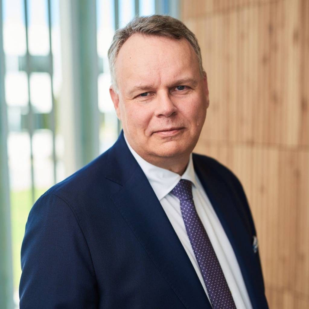 Juho Saari, jolla on lyhyet harmaat hiukset, katsoo kameraan. Hänellä on yllään valkoinen kauluspaita, violetti solmio ja tummansininen pikkutakki.