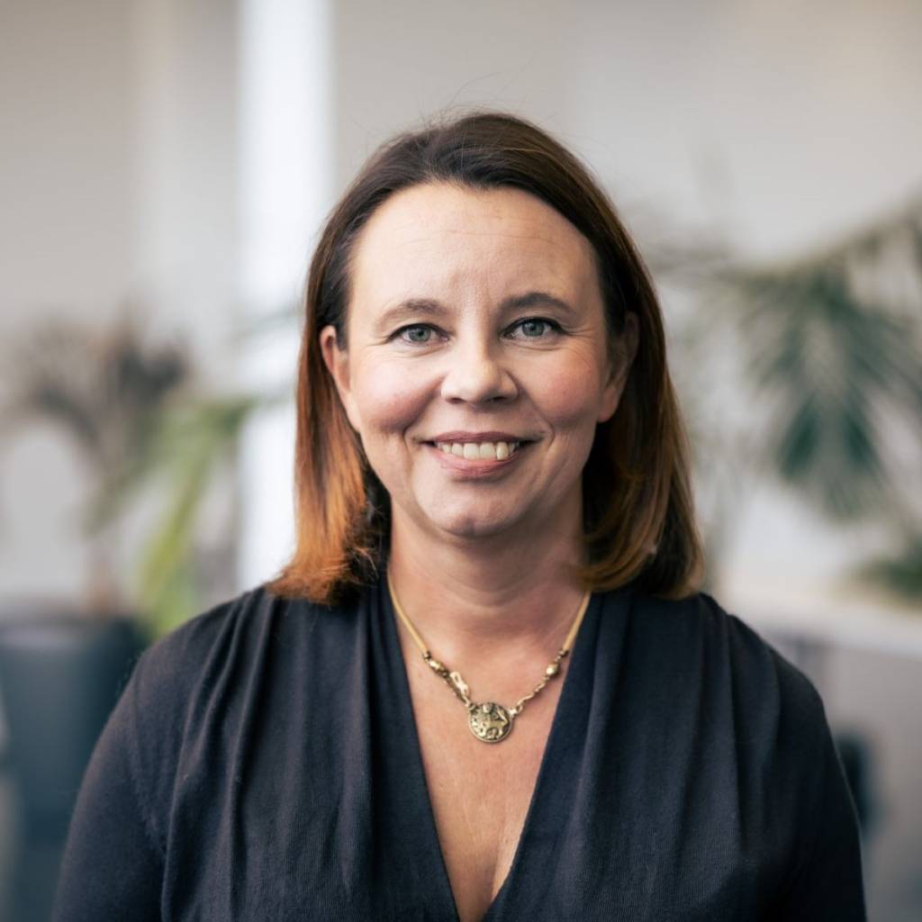 Heli Skottman, jolla on polkkamittaiset punertavanruskeat hiukset, katsoo suoraan kameraan ja hymyilee. Hänellä on musta pusero ja kullanvärinen kaulakoru, jossa riipus. Taustalla näkyy viherkasveja.