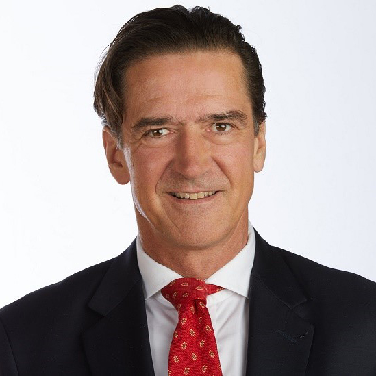 Kai Sauer, jolla on tummat hiukset katsoo kameraan hymyillen. Hän on pukeutunut valkoiseen kauluspaitaan, punaiseen solmioon ja mustaan pikkutakkiin.