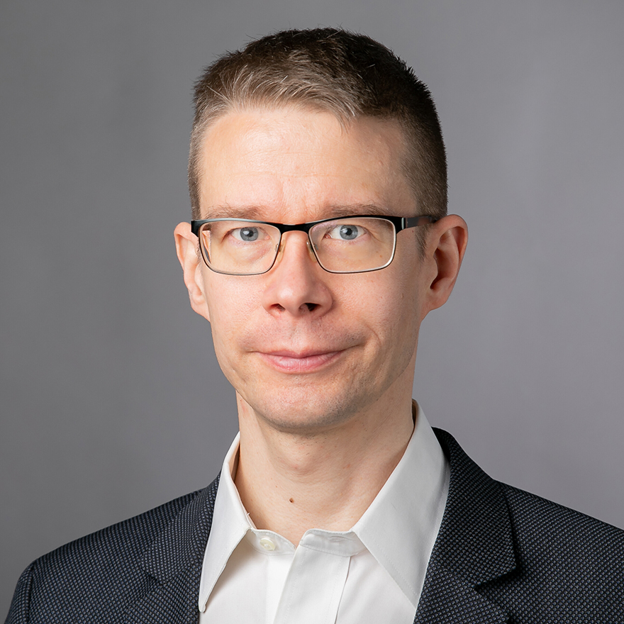 Ville Voipio, jolla on lyhyet tummat hiukset ja silmälasit katsoo kameraan. Hänellä on päällään valkoinen kauluspaita ja tumma pikkutakki.