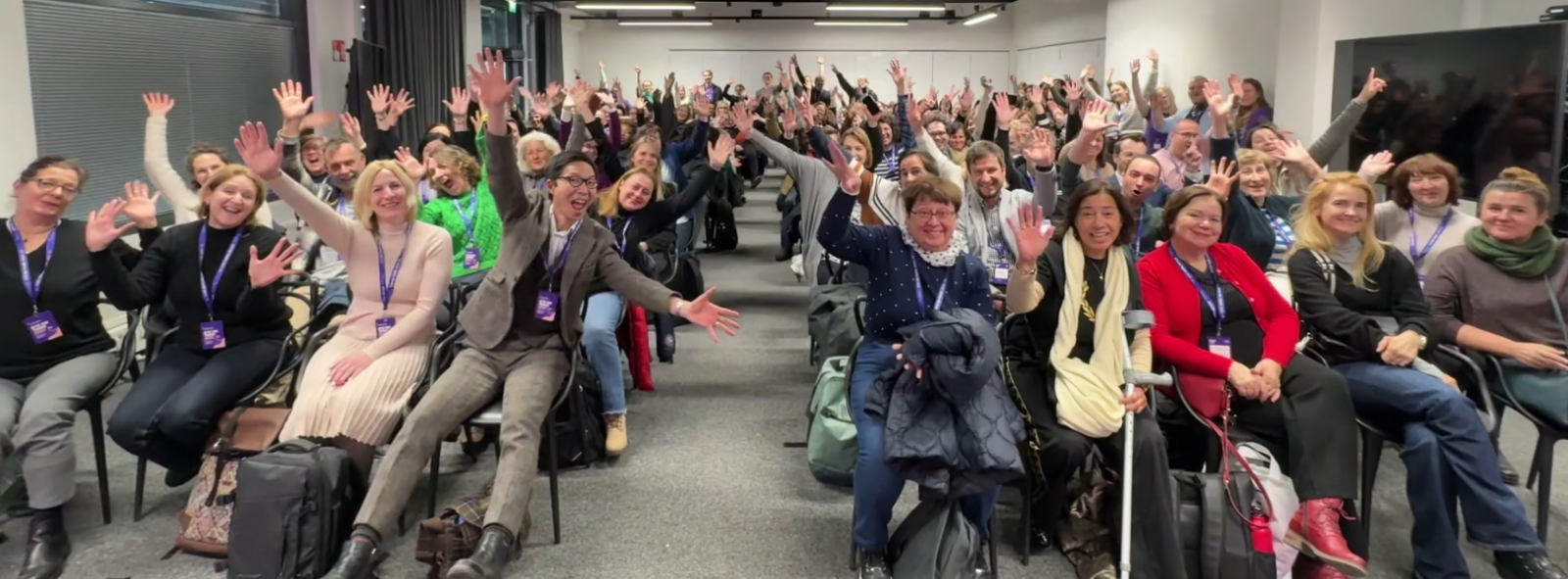 People sitting in a large room, looking at the camera and waving
