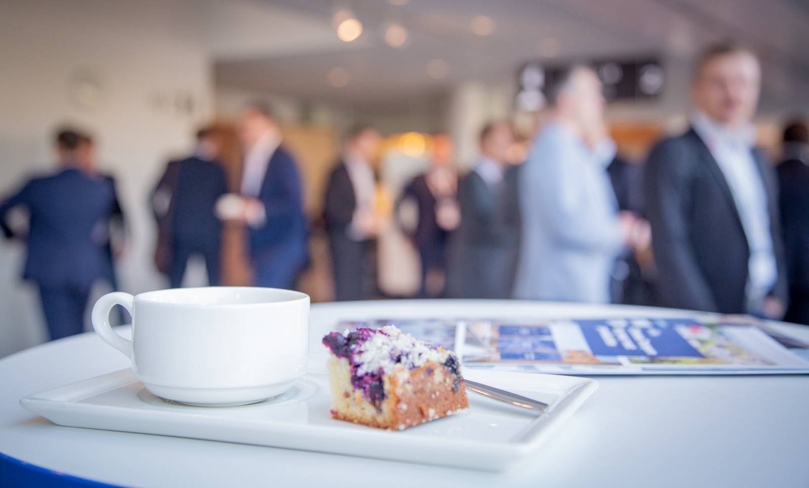 A cup of coffee and a piece of blueberry pie on a table, with conference participants in the background